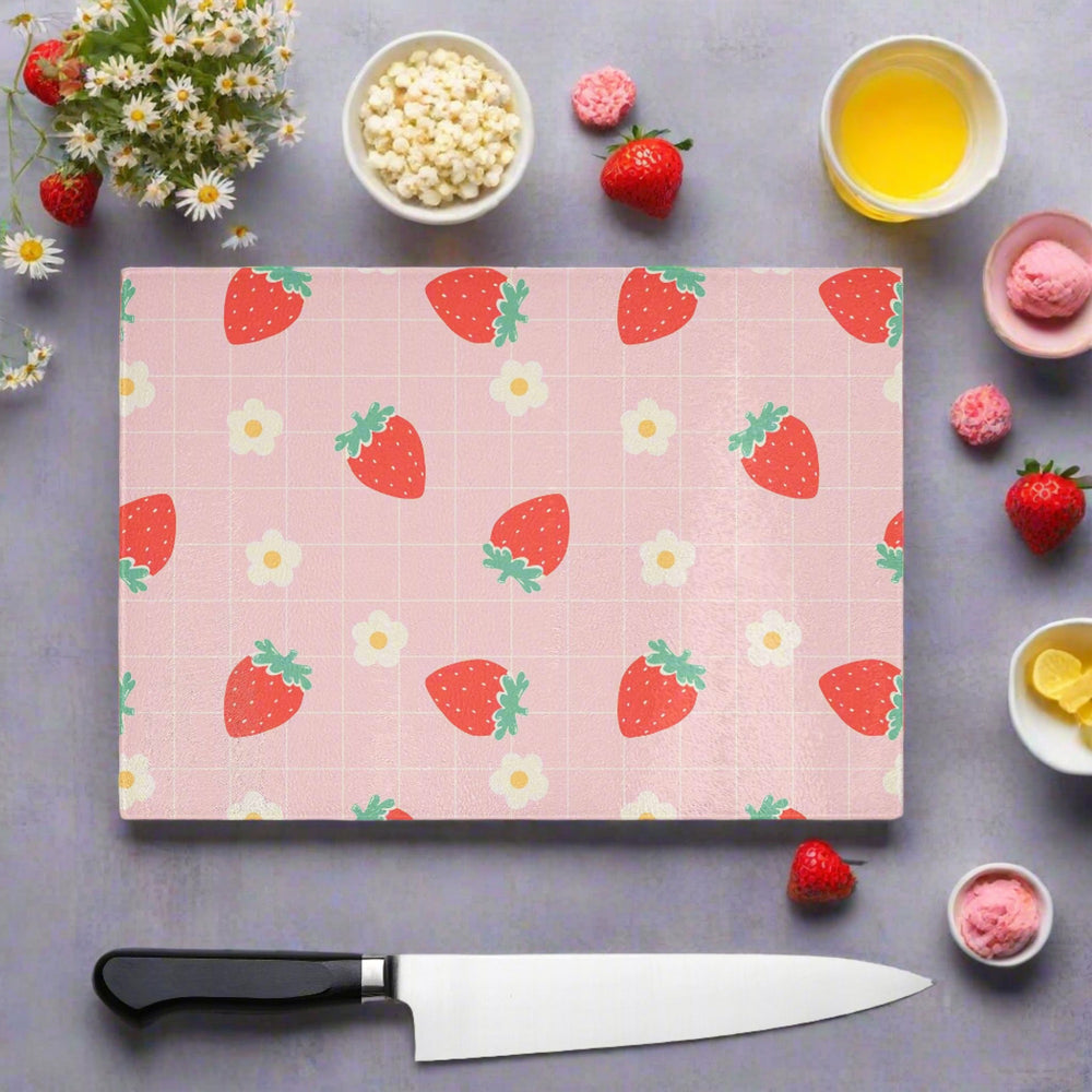 The Yililo Strawberry Check Glass Chopping Board Worktop Saver, in pastel pink cottagecore chic style with a strawberry pattern, is shown on a gray surface amid a knife, strawberries, lemon slices, flowers, popcorn, juice, and ice cream.
