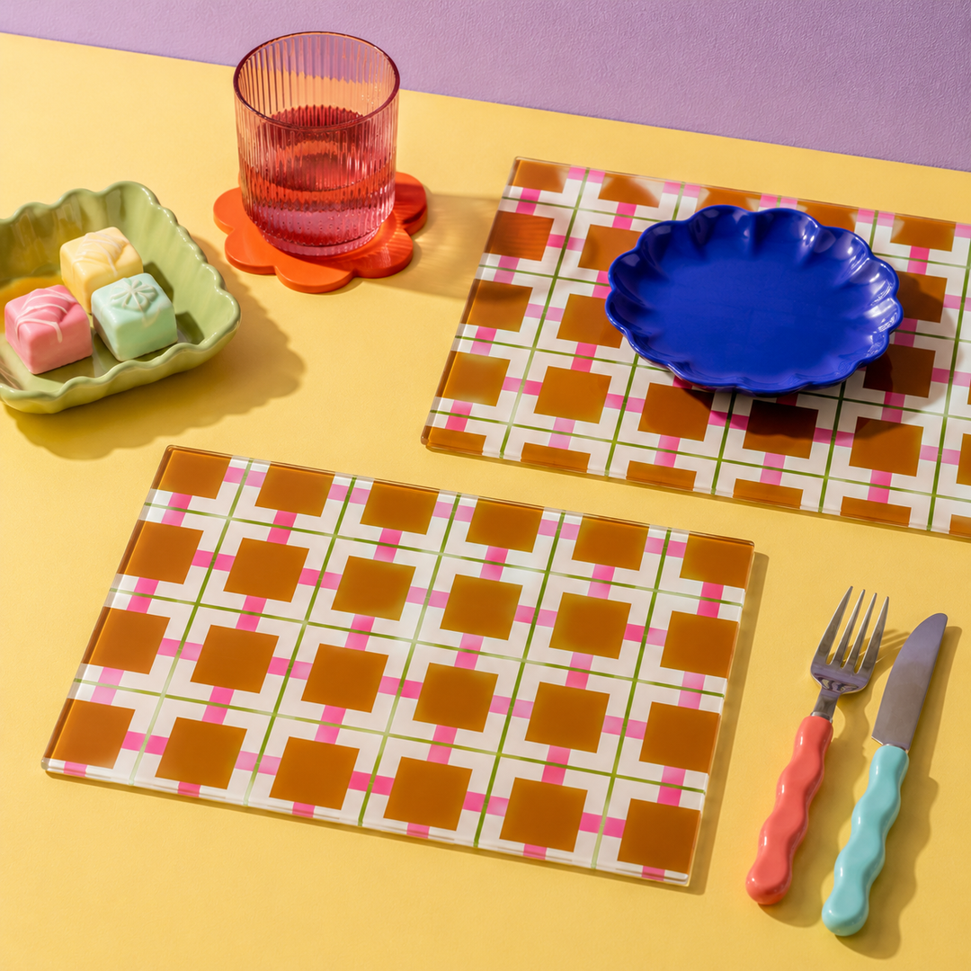 A styled tabletop scene with two glass rectangular placemats featuring a geometric check pattern in brown, pink, and white, alongside a blue scalloped plate, a pink ribbed glass, a small dish with pastel cubes, and a pink-and-blue utensil set on a yellow surface.