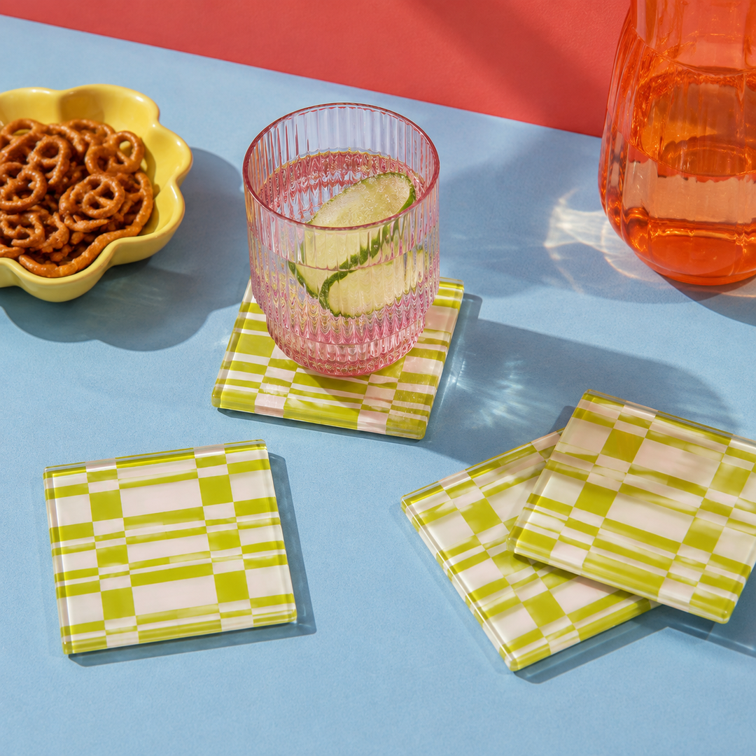 Green and white checkered square coasters displayed on a light blue table with a pink glass, pretzels in a yellow bowl, and an orange pitcher in the background.