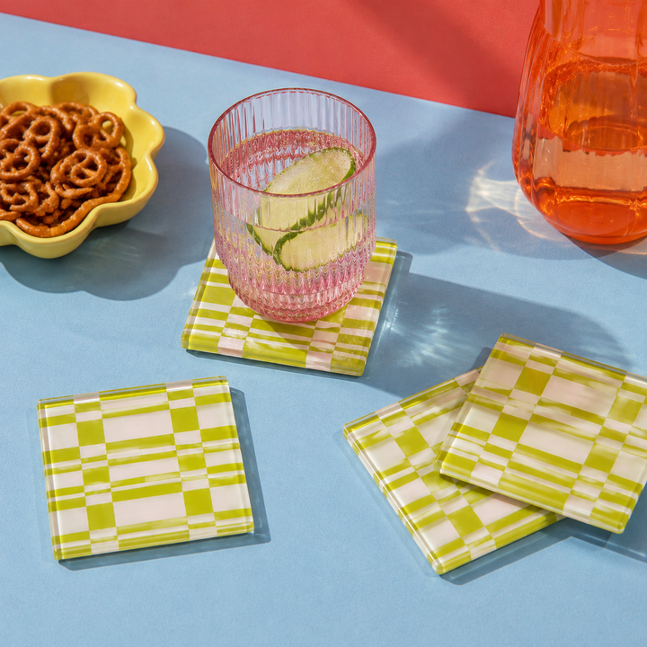 Green and white checkered square coasters displayed on a light blue table with a pink glass, pretzels in a yellow bowl, and an orange pitcher in the background.