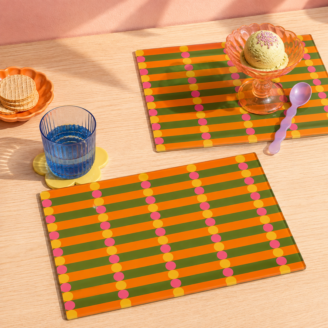 Two rectangular glass placemats with horizontal orange, green, and yellow stripes and pink and yellow dots, styled on a light wood table with a glass of water, cookies, a dessert in a glass, and a purple spoon.