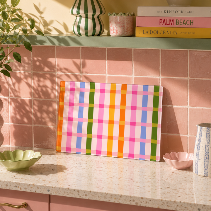 A colourful plaid cutting board with vertical stripes in orange, yellow, green, blue, pink, and white, displayed on a speckled kitchen counter against a pink tiled backsplash.