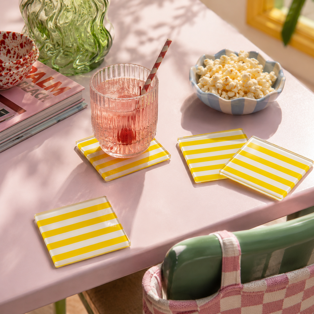 Four square yellow and white striped glass coasters arranged on a pink table, with a pink drink, popcorn bowl, and other lifestyle props in a sunlit indoor setting.