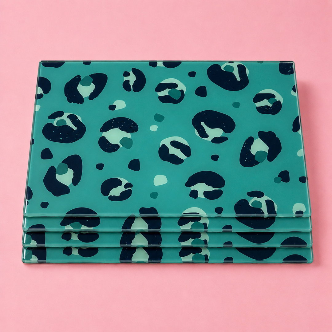 A stack of rectangular glass tablemats with a teal background and a dark navy and mint green leopard print pattern, displayed on a solid pink background.