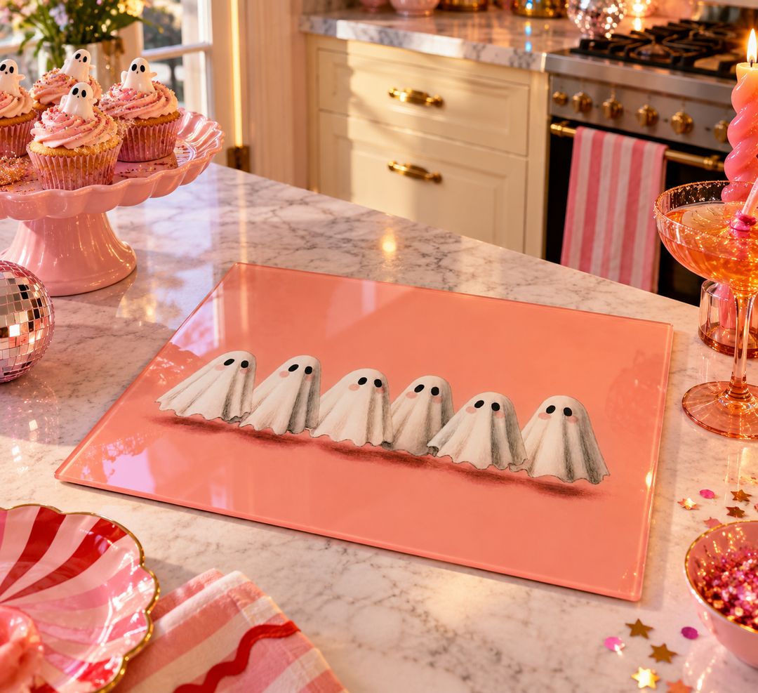Six white ghost-shaped cookies with black eyes are arranged on a glossy coral pink rectangular serving board on a marble kitchen counter. Pink party decorations, cupcakes, and a candle are visible in a decorated kitchen setting.