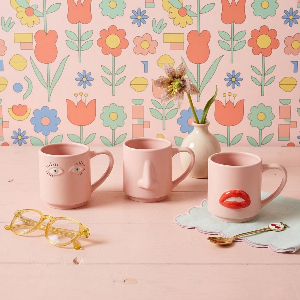 Three pink mugs with face designs on a table with a floral wallpaper background.