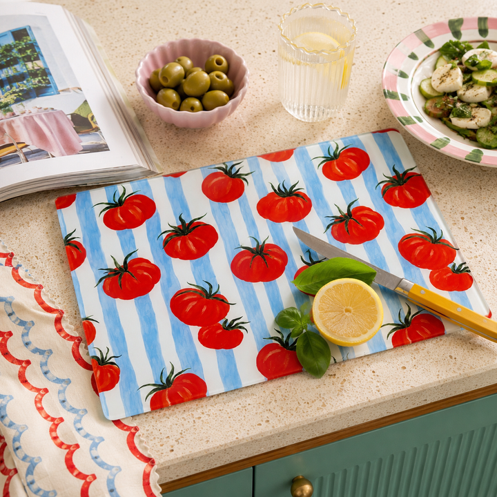 A rectangular kitchen cutting board with a blue and white striped background and a red tomato print, styled on a beige countertop with a knife, lemon, olives, and a plate of food nearby by yililo 