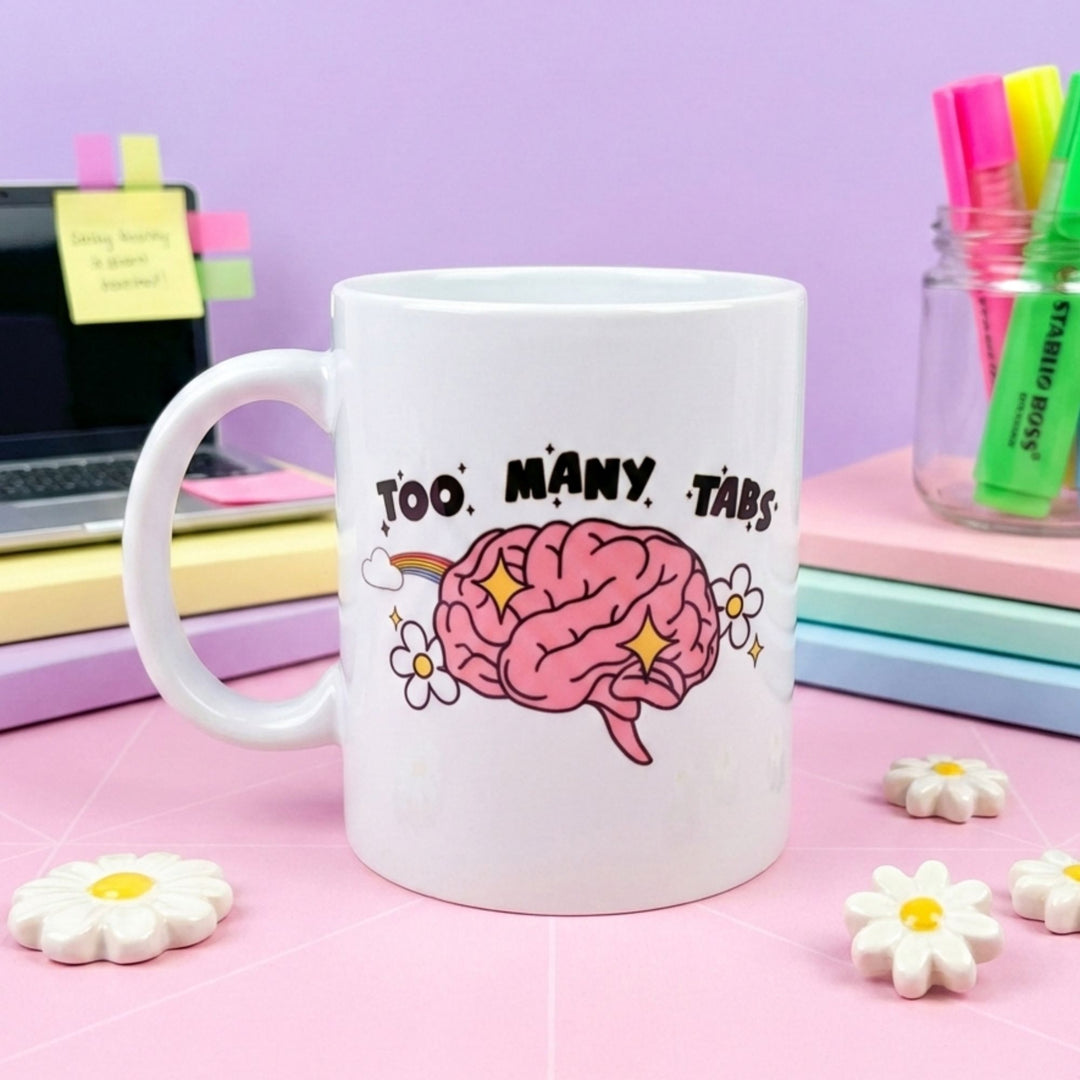 White mug with a brain graphic and 'Too Many Tabs' text on a pink surface with flowers and stationery.