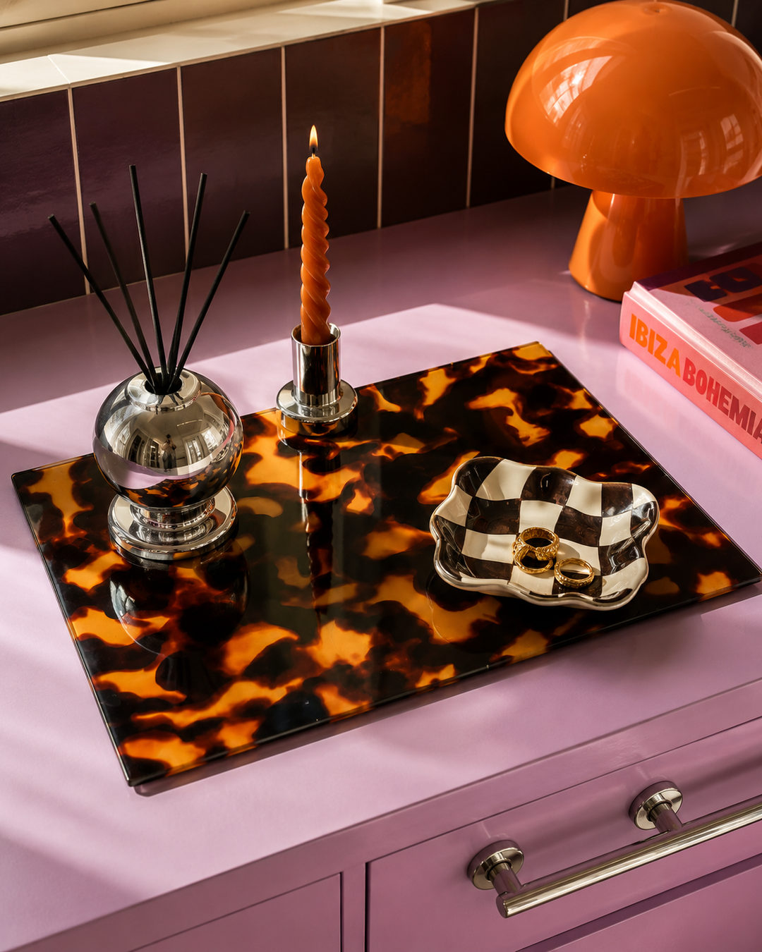 A glossy tortoiseshell glass worktop saver being used as a display tray with a black, orange, and brown pattern sits on a lavender-purple dresser. The tray is styled with a silver reed diffuser, a twisted orange candle, a checkered black-and-white dish, and a pink book.