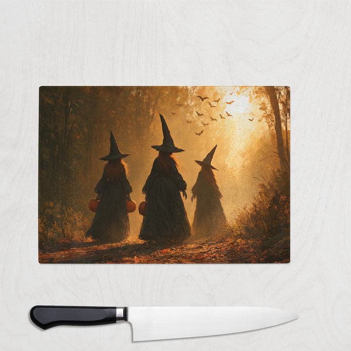 Trick Or Treat Witches glass chopping board with three witches carrying pumpkin buckets through a golden autumn wood