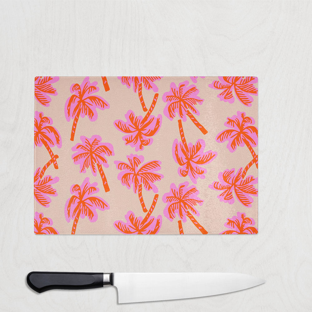 The Yililo Tropical Vibes Glass Chopping Board, featuring a pink and orange palm tree design, rests on a light wooden surface with a chef’s knife placed below.