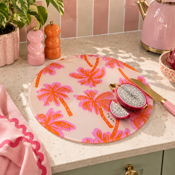 A round glass cutting board with a pink and orange palm tree pattern, styled on a kitchen counter with dragon fruit, a pink towel, and kitchen accessories.