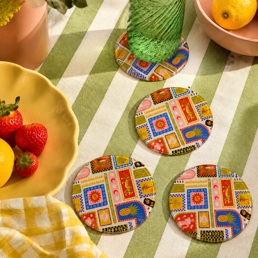 Colorful patterned glass coasters on a green and white striped tablecloth with a glass of green liquid and fruit.