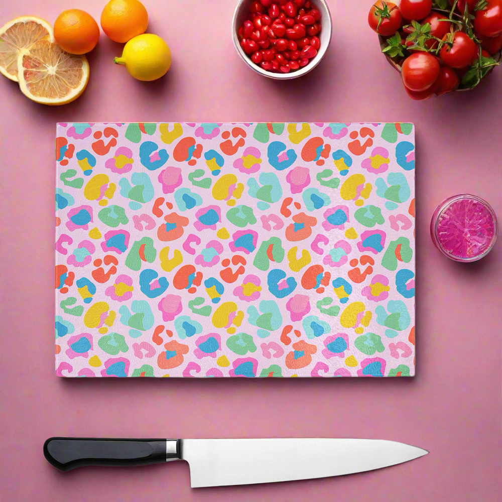 The Yililo Wild Rainbow Leopard Print Glass Chopping Board features vibrant abstract patterns, displayed on a pink surface with a knife, sliced lemon, cherry tomatoes, red berries, an orange, and a glass of pink drink. Perfect for colourful kitchens.