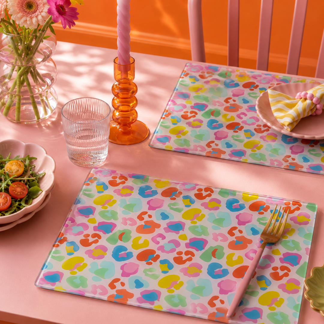 Two rectangular placemats with a colorful pastel abstract pattern are styled on a pink table, surrounded by a glass, candle, flowers, and plates in a bright, warm setting by yililo