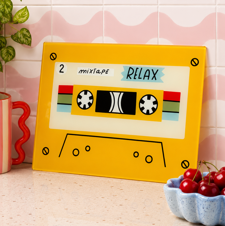 Yellow tape designed colourful glass chopping board leaned against tiles with a bowl of cherries in front 