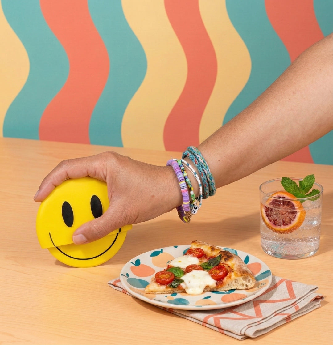 Hand holding a smiley face object with a plate of pizza and a glass of water on a colorful background