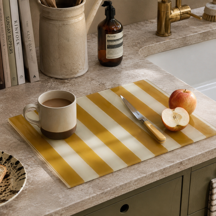 A rectangular glass cutting board with bold yellow and white diagonal stripes sits on a stone countertop in a kitchen setting. The board is styled with a ceramic mug, a knife, a whole apple, and a halved apple.