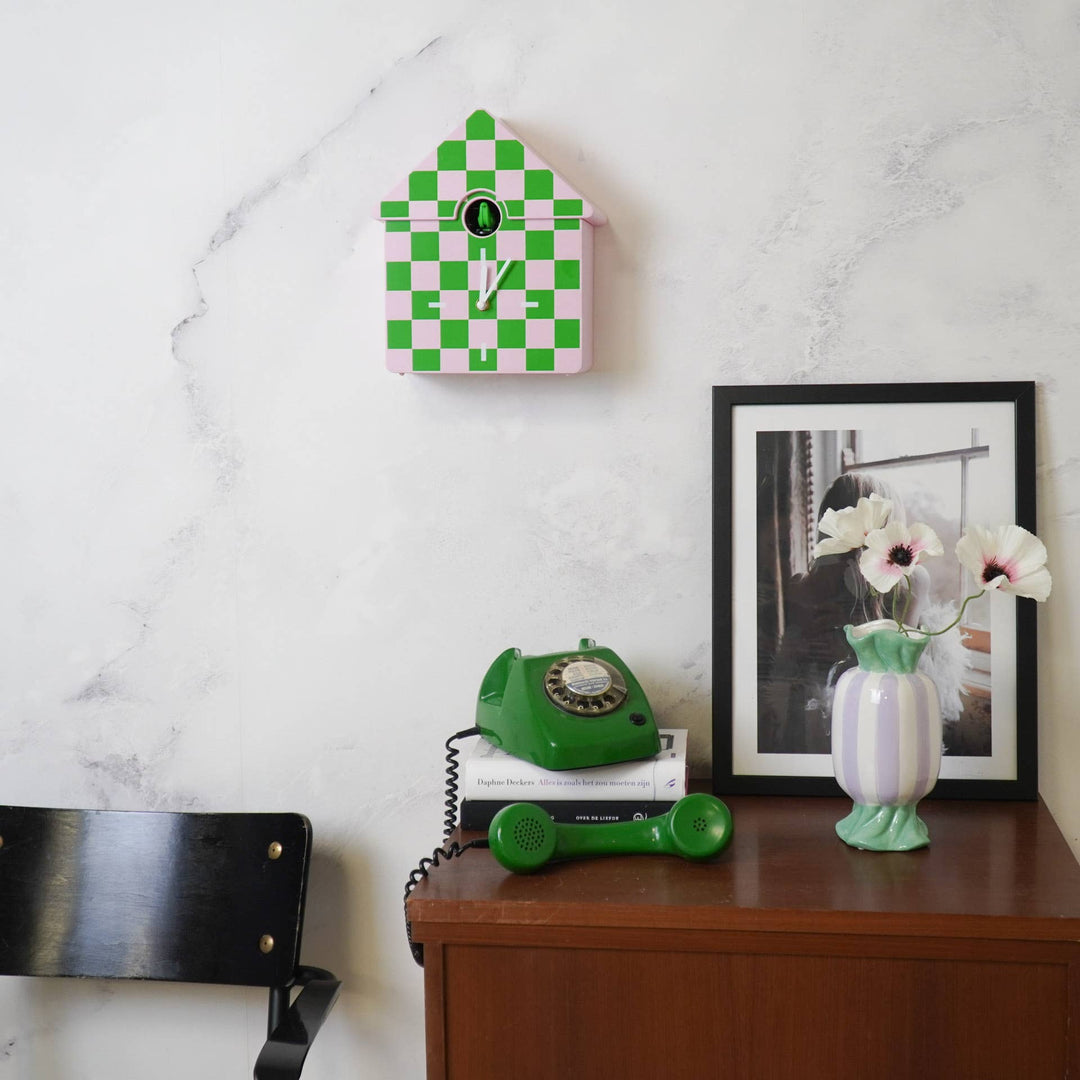 Green checkered clock on a white wall above a wooden desk with a green phone and vase.