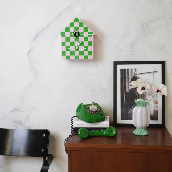 Green checkered clock on a white wall above a wooden desk with a green phone and vase.