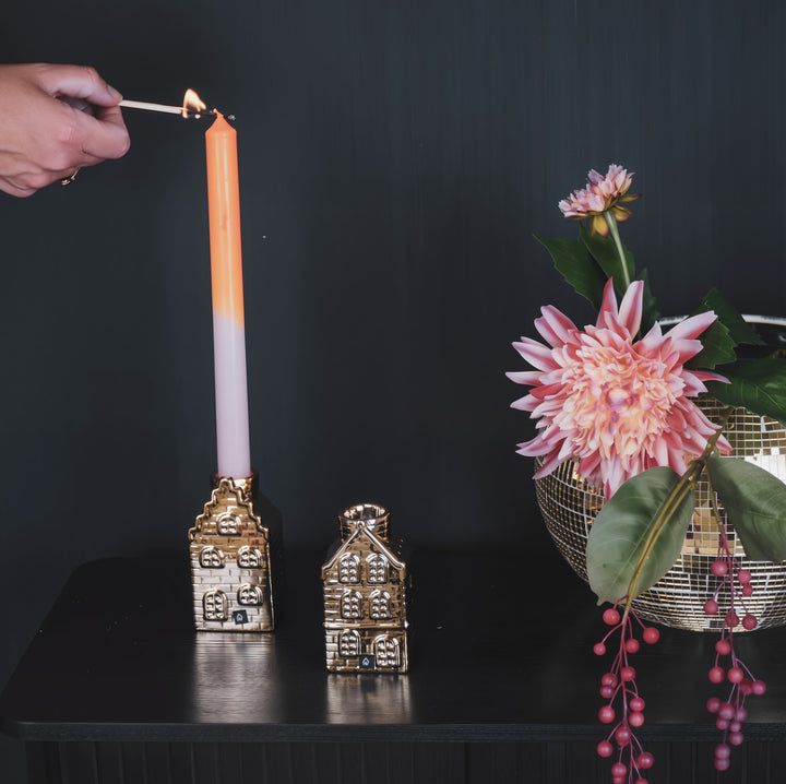 Candle being lit with decorative gold house candle holders and a floral arrangement on a dark background