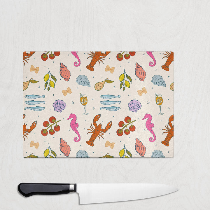 Colourful repeat print of lobsters, lemons, Aperol spritz glasses, seahorses and pasta on a glass chopping board – summery and fun kitchen accessory.