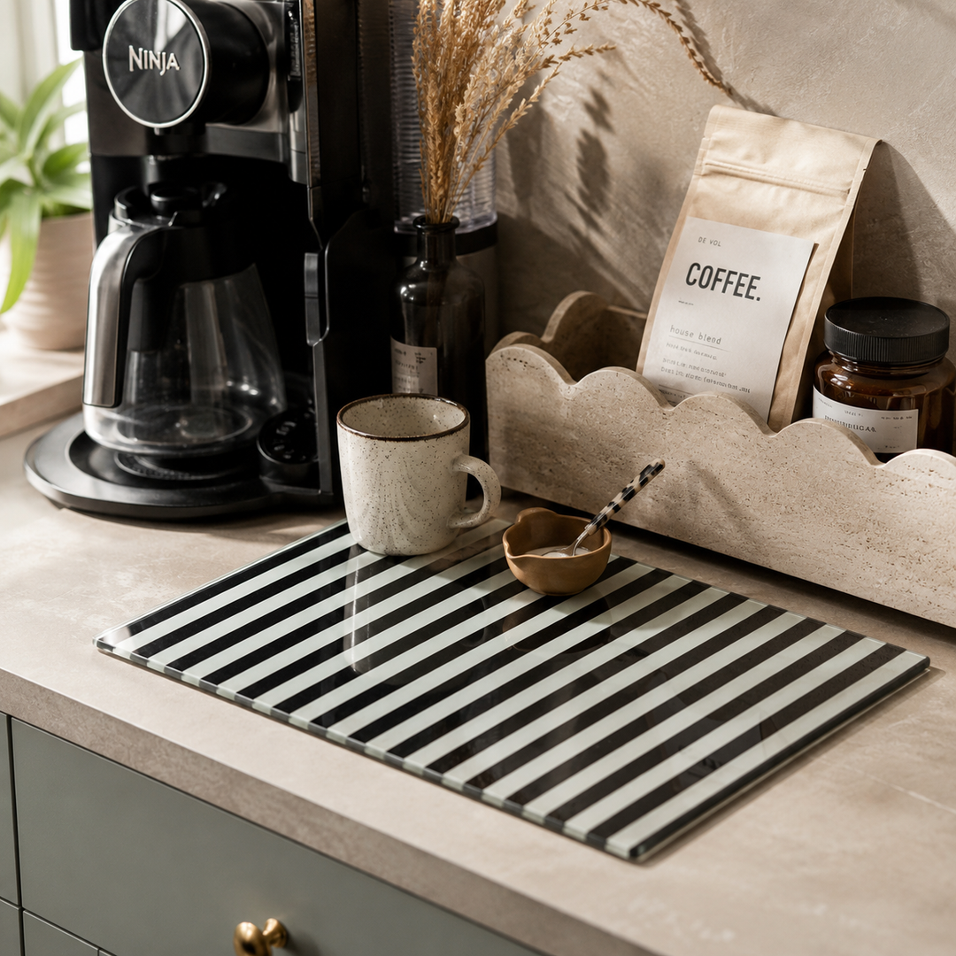 A rectangular black and white striped glass serving board on a beige countertop, styled with a speckled mug, a small bowl, and a coffee bag in a kitchen setting.