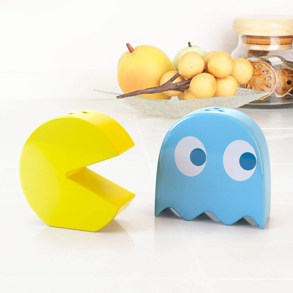 Two Pac-Man ghost-shaped objects salt and pepper shakers, one yellow and one blue, on a white surface with fruit in the background.