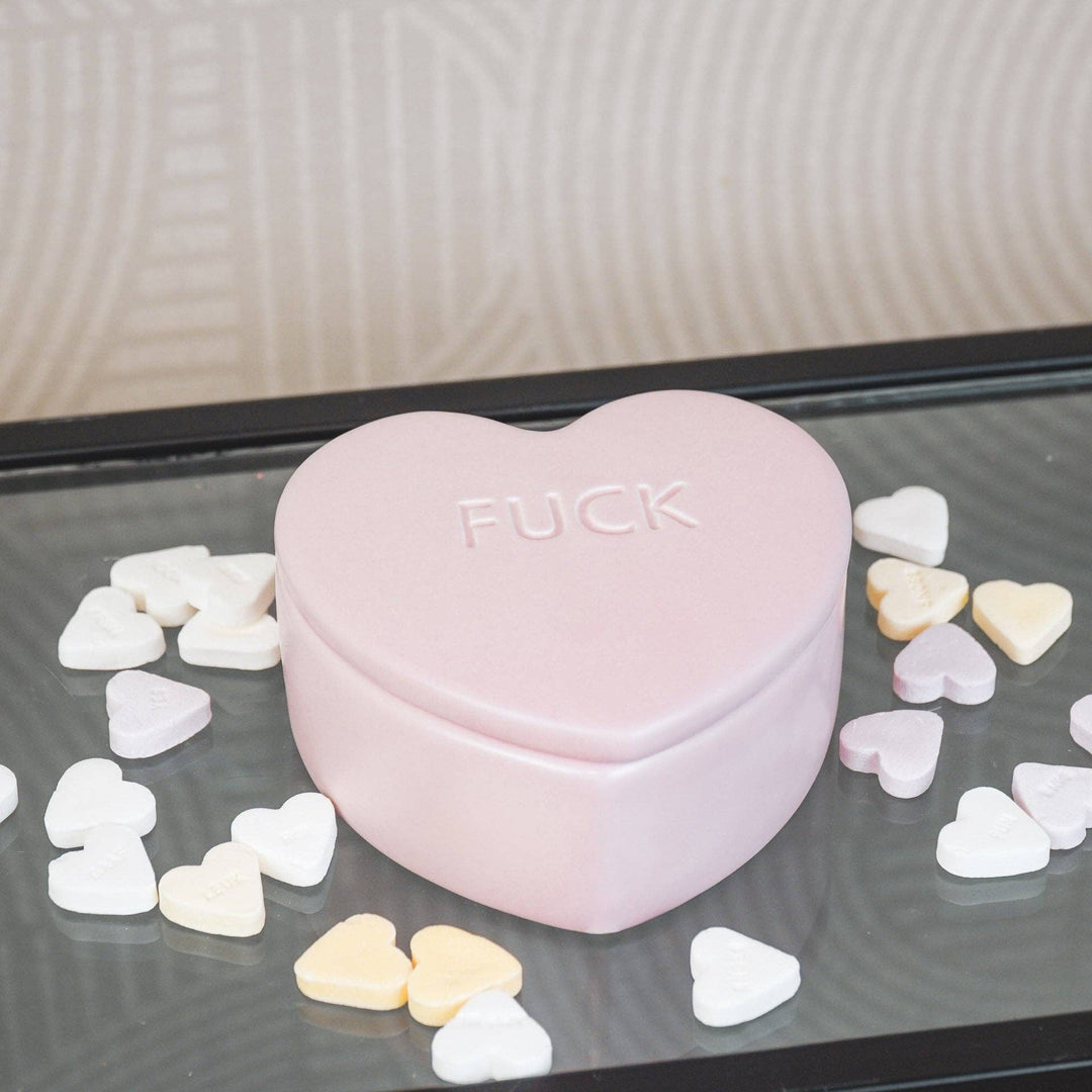 Heart-shaped pink container with 'FUCK' text surrounded by small heart-shaped objects on a reflective surface.