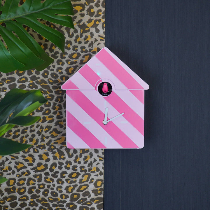 Pink and white striped cuckoo clock on a dark surface with a patterned fabric and leaves in the background