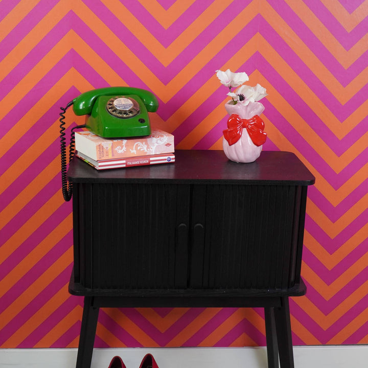 Vintage green phone and decorative vase on a black cabinet against a pink and orange chevron wall.