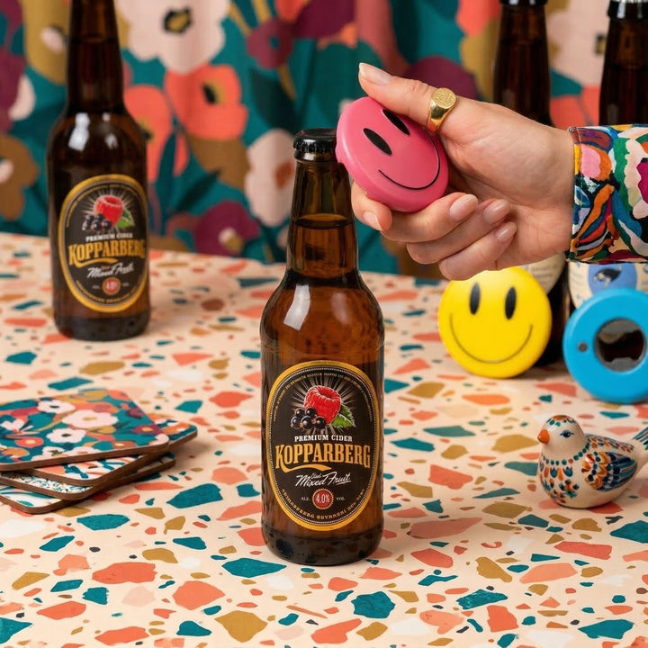 Kopparberg cider bottle with a hand holding a pink smiley face object on a colorful table.