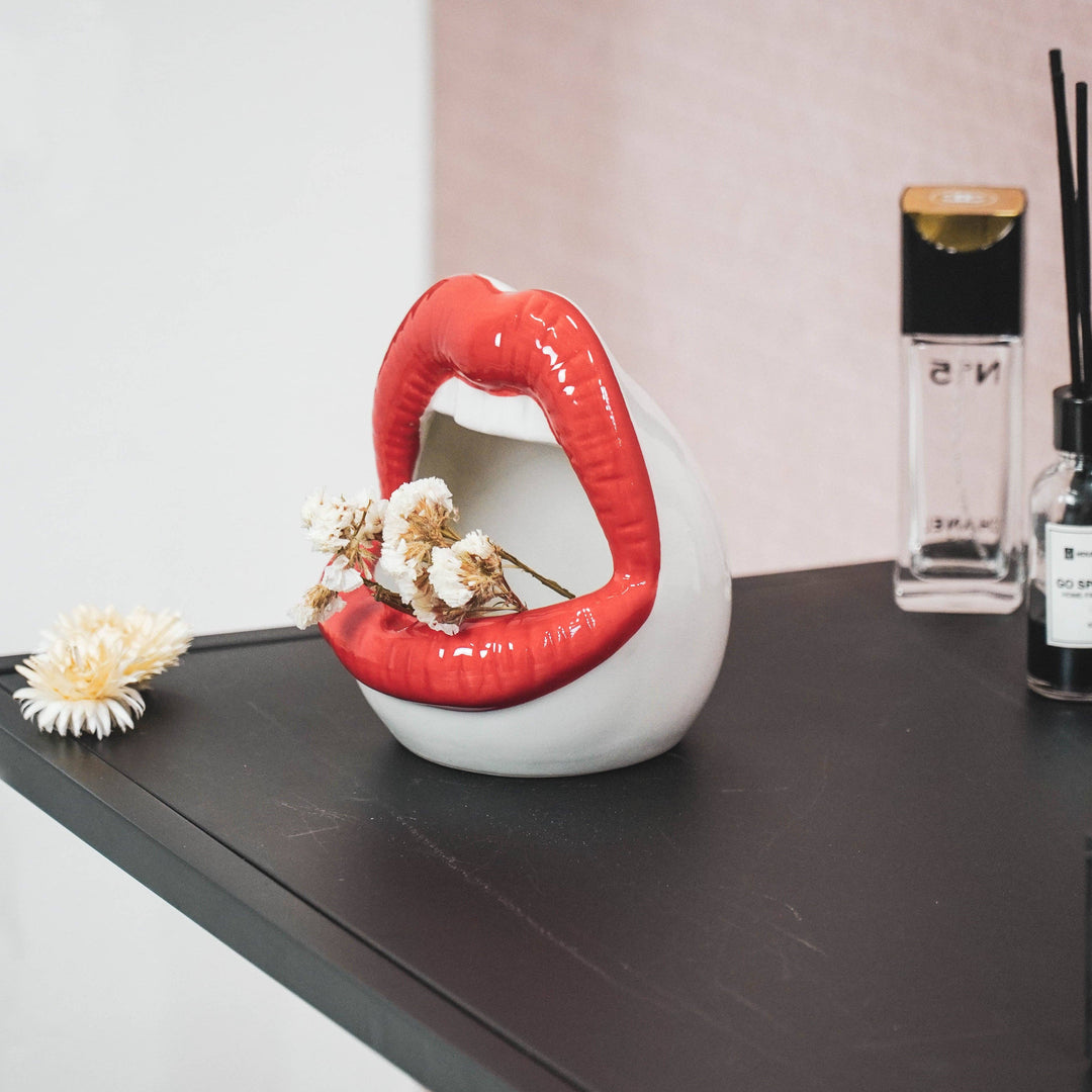 Lip-shaped vase with red lips and white body on a dark surface with flowers and perfume bottles.