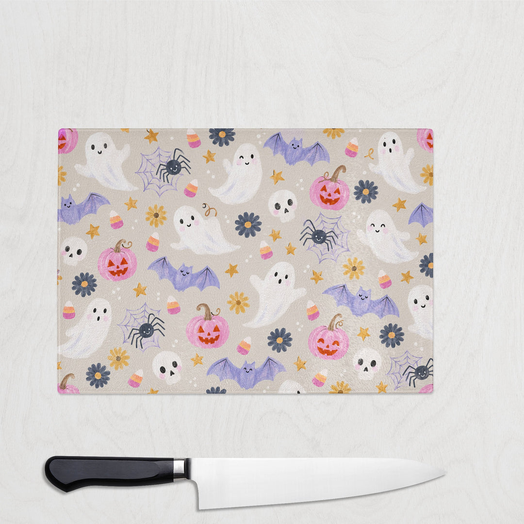 Cutesy Pink Halloween glass chopping board on a white wood worktop with a chef's knife, smiling ghosts, lilac bats and pink pumpkins on soft oatmeal