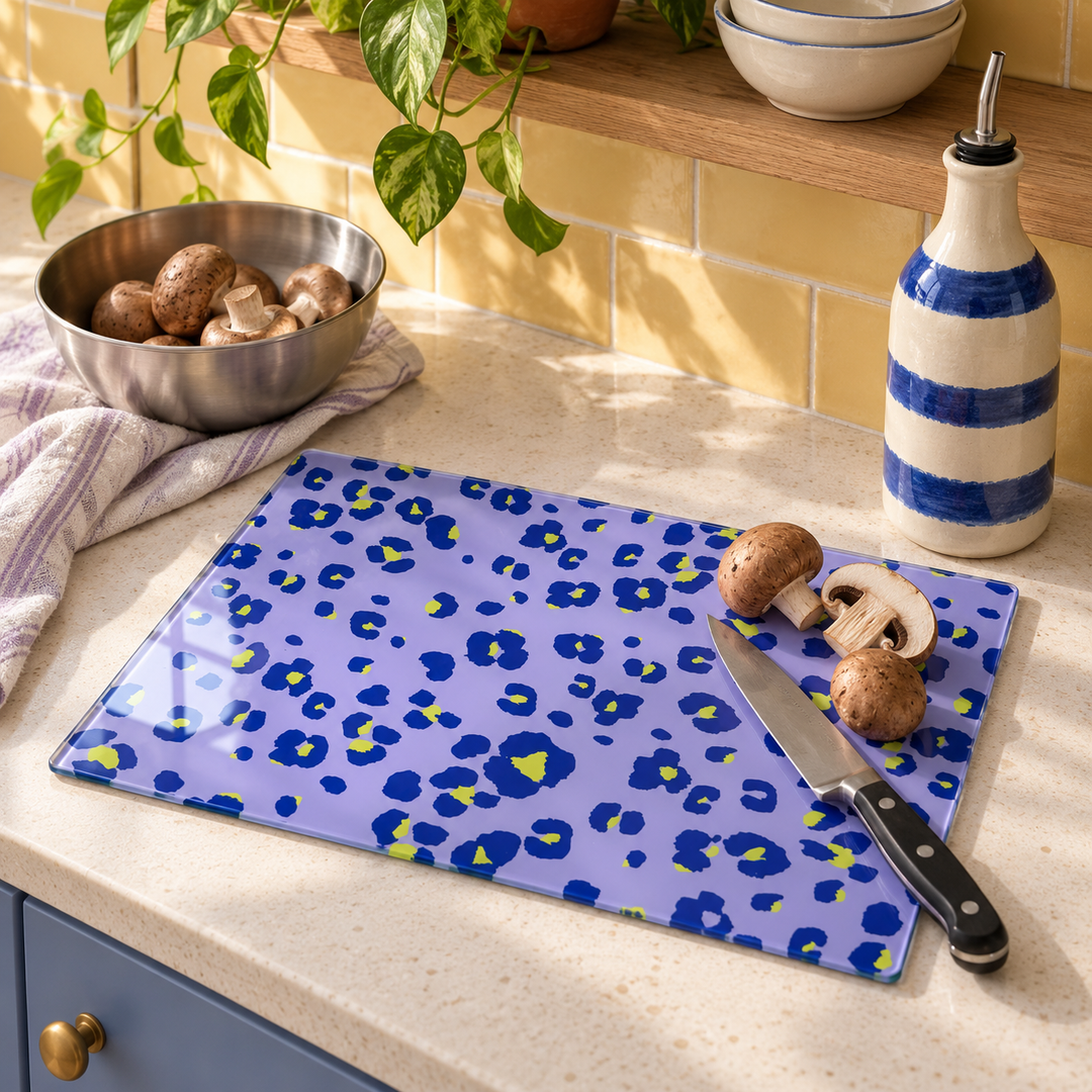 Lilac Lime Neon Leopard glass chopping board on a kitchen worktop with chestnut mushrooms, a chef's knife and a blue striped oil bottle, yellow tiles behind