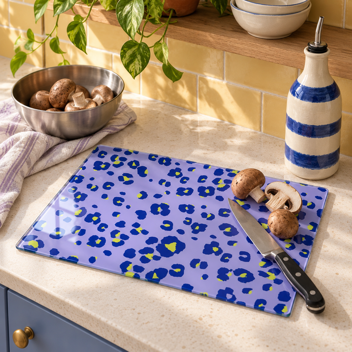Lilac Lime Neon Leopard glass chopping board on a kitchen worktop with chestnut mushrooms, a chef's knife and a blue striped oil bottle, yellow tiles behind