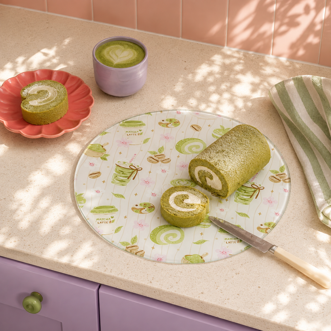 Matcha and Cake round glass chopping board on a cream worktop with a sliced matcha swiss roll, a matcha latte in a lilac mug and a pink scalloped plate, pink tiles behind