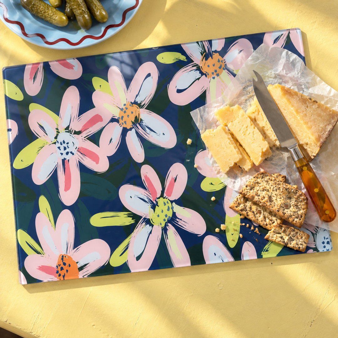 Midnight Bloom glass chopping board used as a cheese board on a yellow table with cheddar, seeded crackers, a cheese knife and a plate of gherkins