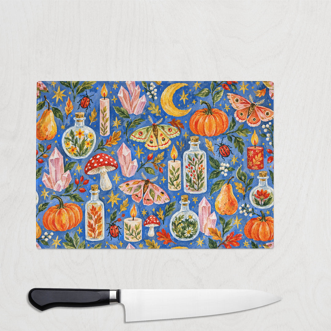 Practically Magic glass chopping board on a white wood worktop with a chef's knife, pumpkins, potion bottles, crystals, moths and candles painted on rich blue