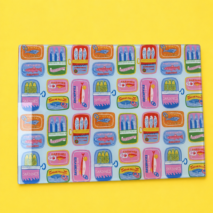 Sardine Parade glass chopping board with colourful illustrated sardine tins, laid flat on a bright yellow background