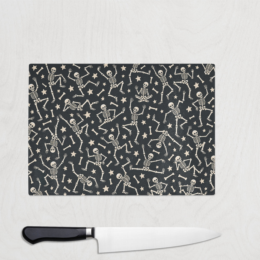 The Little Skeletons glass chopping board on a white wood worktop with a chef's knife, a crowd of tiny dancing skeletons and stars on near-black