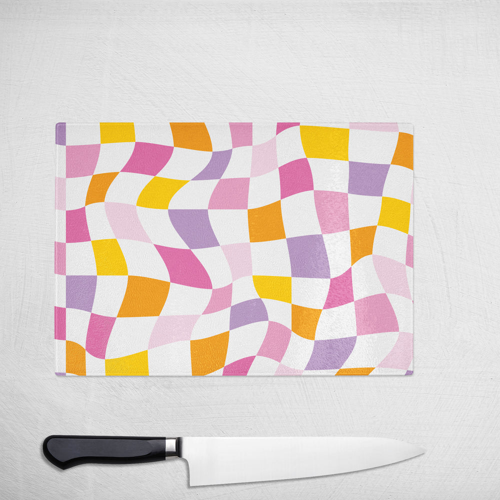 The Yililo Colourful Wavy Check Glass Chopping Board, featuring bold pink, purple, orange, and yellow patterns, adds stylish flair to any countertop—shown here on a white surface with a large kitchen knife below it.