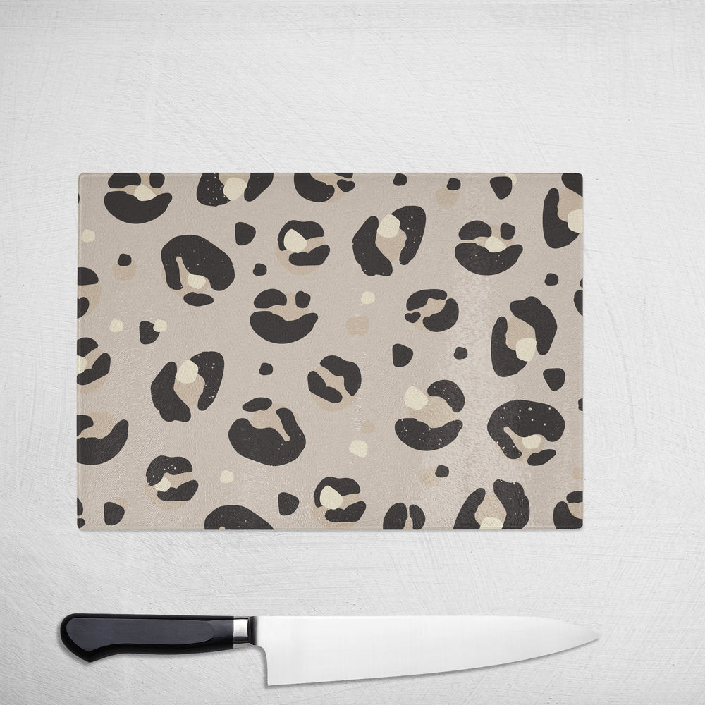 The Yililo Cream Leopard Print Glass Chopping Board features a beige animal-print design and rests on a white textured surface, paired with a black-handled kitchen knife below it.