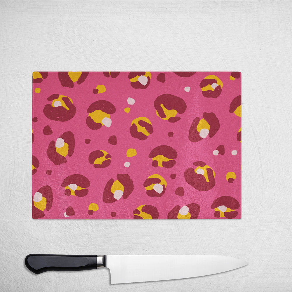 The Yililo Dark Pink And Yellow Glass Chopping Board, featuring an abstract pattern, rests on a white textured surface—a stylish kitchen essential and perfect gift for cooks. Includes a sleek kitchen knife below the board.