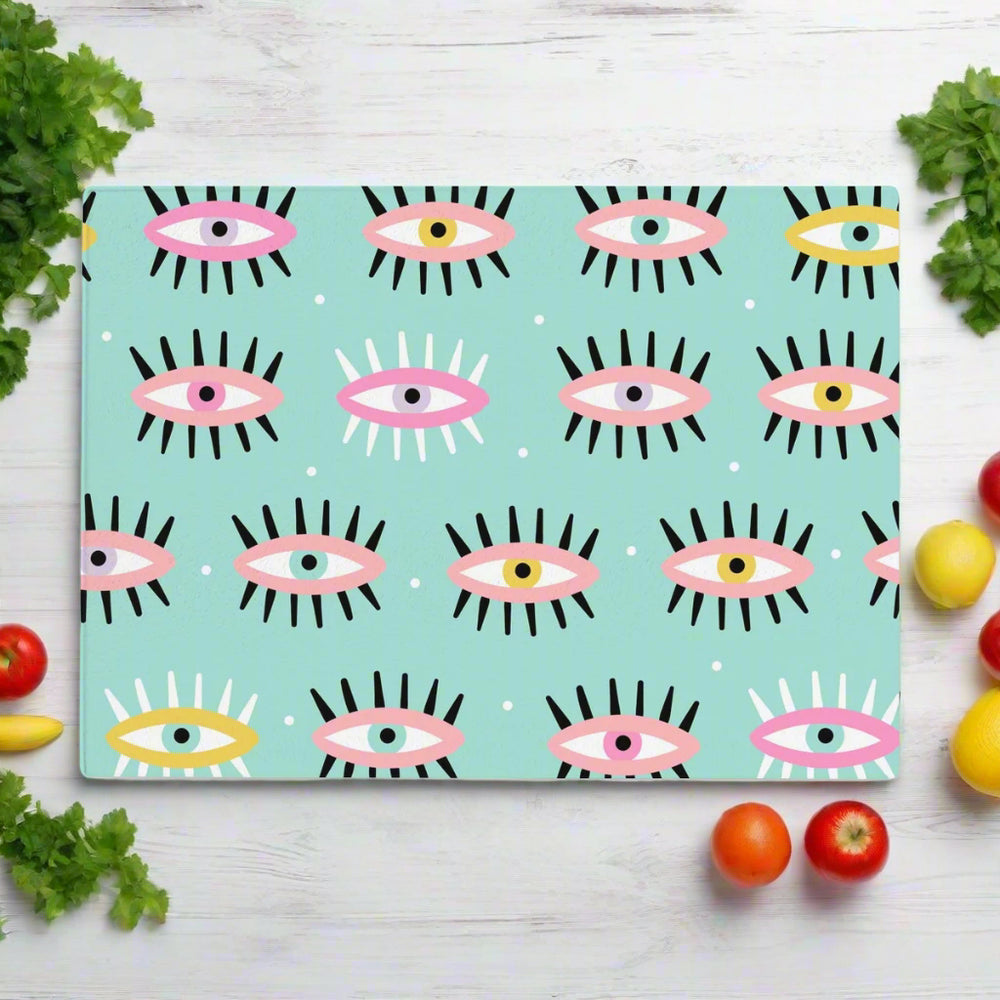 The Yililo Evil Eye Green Glass Chopping Board features a whimsical cartoon eye pattern in pastel blue green—an ideal tempered glass gift for home cooks, shown surrounded by fresh vegetables on a white wooden surface.
