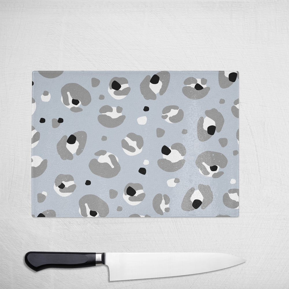 The Yililo Grey Leopard Print Glass Chopping Board, featuring an abstract blue, white, black, and gray design, sits on a textured white surface beside a black-handled chefs knife—a stylish gift for any home chef.