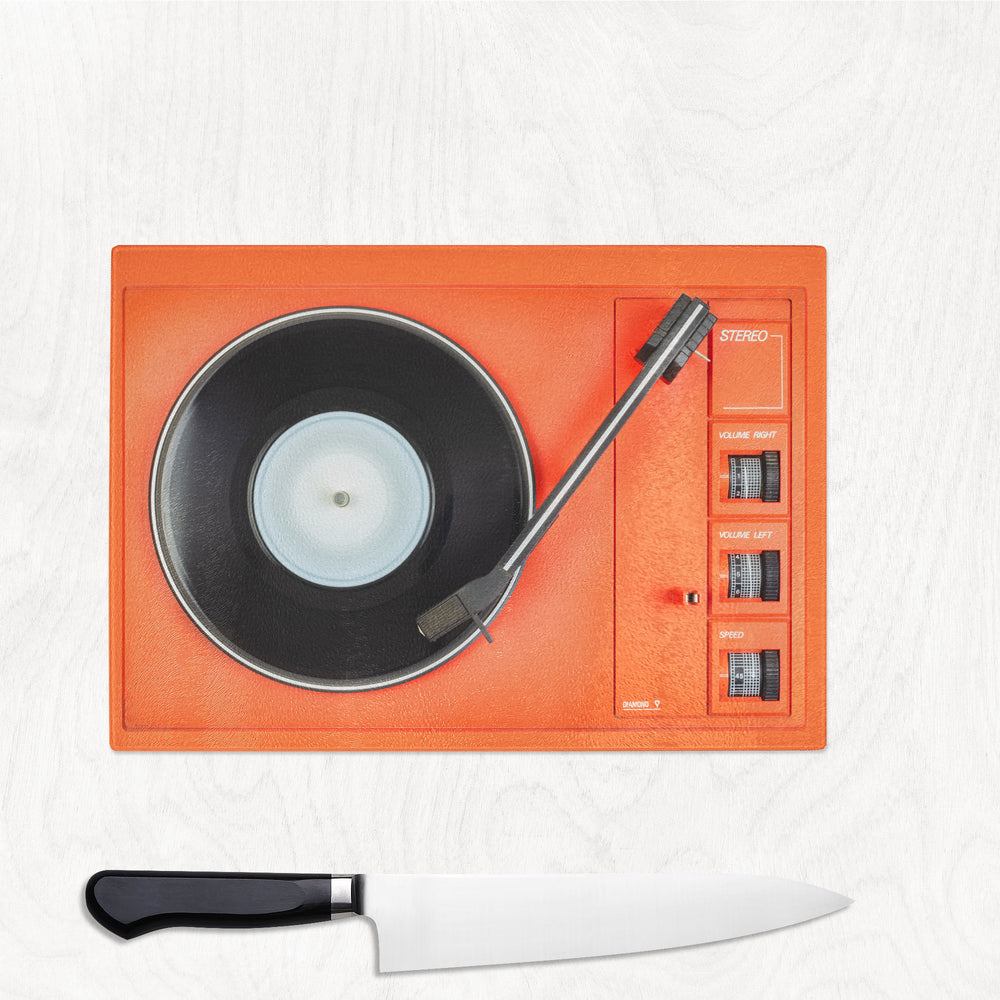 The Yililo Orange Record Player Turntable Glass Chopping Board rests on a light wooden surface, paired with a black-handled kitchen knife—an ideal retro gift for music lovers and stylish homes.