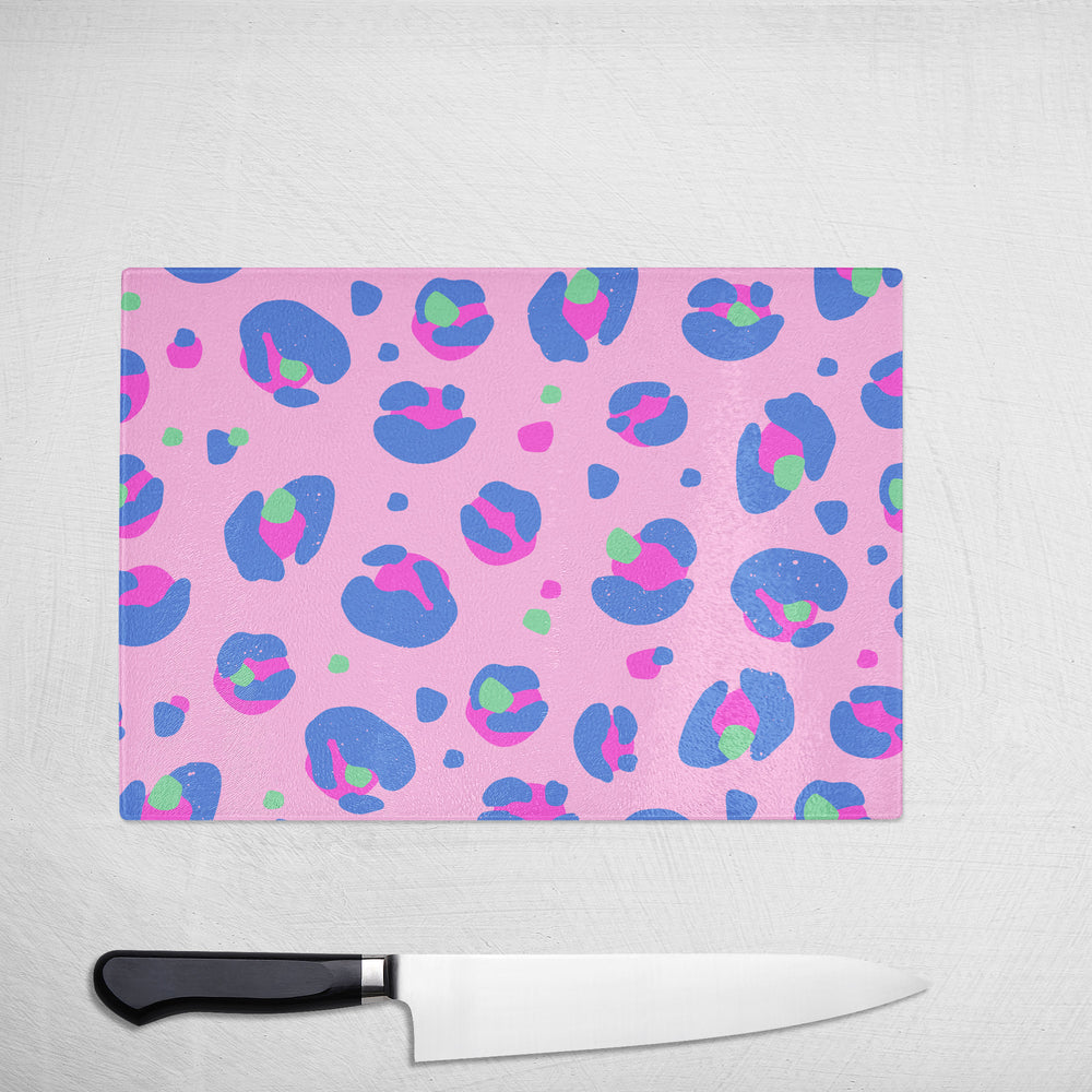 The Yililo Pink Blue Leopard Print Glass Chopping Board features abstract spots and makes a stylish kitchen gift, shown here beside a chefs knife on a white textured surface.