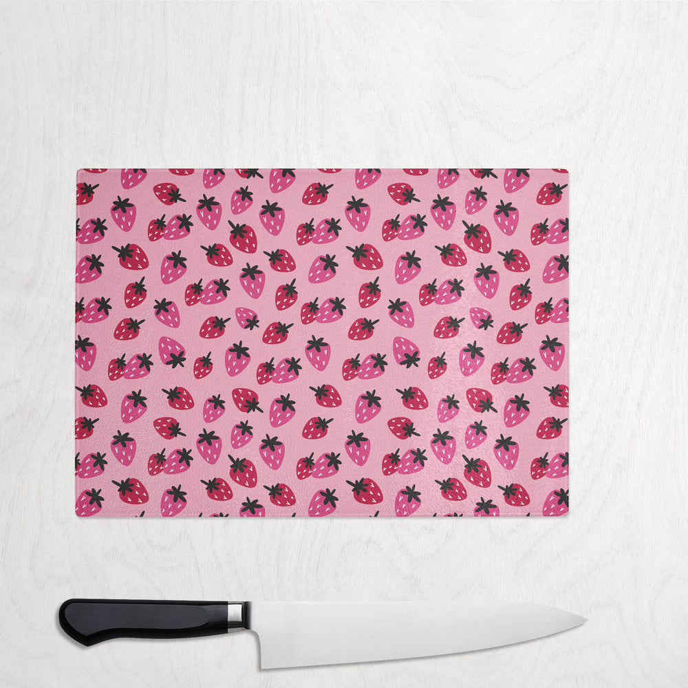 The Yililo Pink Strawberry Glass Chopping Board Worktop Saver, featuring red and black berry prints on a pink background, rests on a light wood surface with a chef’s knife with a black handle placed below it.